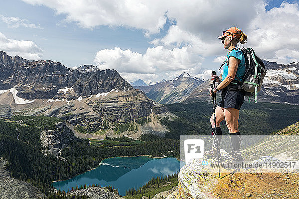 Female hiker standing on cliff edge overlooking mountains and valley with colourful alpine lake; British Columbia  Canada
