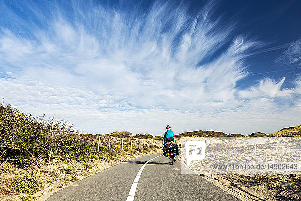 Radfahrerin auf gepflastertem Radweg entlang hügeliger Sanddünen mit dramatischen Wolken und blauem Himmel  südlich von Zandvoort; Niederlande