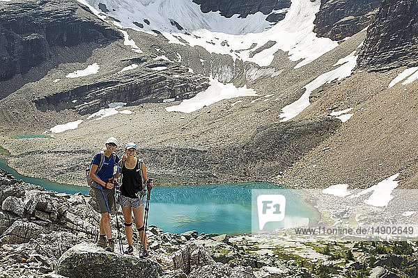 Two female hikers standing in a large rocky area with a colourful alpine lake and mountain cliffs with snow in the background; British Columbia  Canada