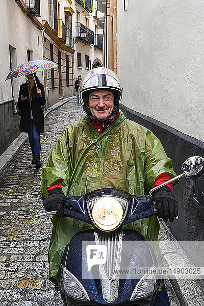 Ein Mann  der einen Poncho und einen Helm trägt  sitzt auf einem Motorroller in einer engen Straße und posiert für die Kamera; Sevilla  Spanien