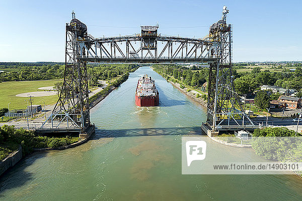 Aerial view of large laker ship navigating under a metal lift bridge in a canal with blue sky; Thorold Ontario Canada