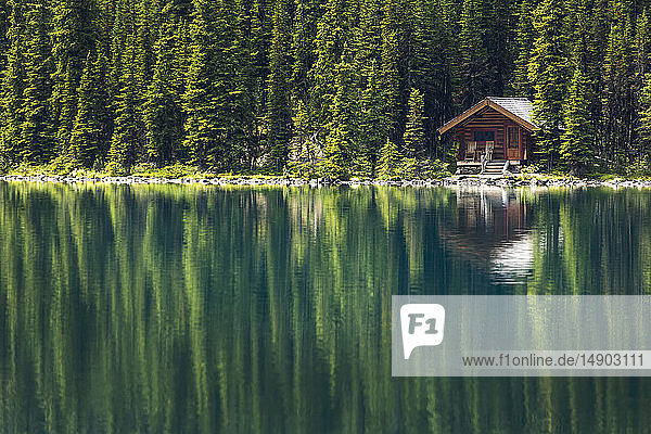 Blockhaus zwischen immergrünen Bäumen an einem alpinen Seeufer mit sanfter Reflexion; British Columbia  Kanada