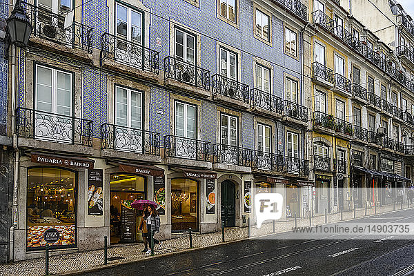 Shops and apartments along a street with pedestrians holding an umbrella on a rainy day; Lisbon  Lisboa Region  Portugal