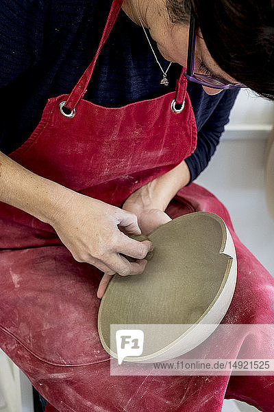 Frau mit roter Schürze sitzt in ihrer Keramikwerkstatt und arbeitet an einer Tonschüssel.