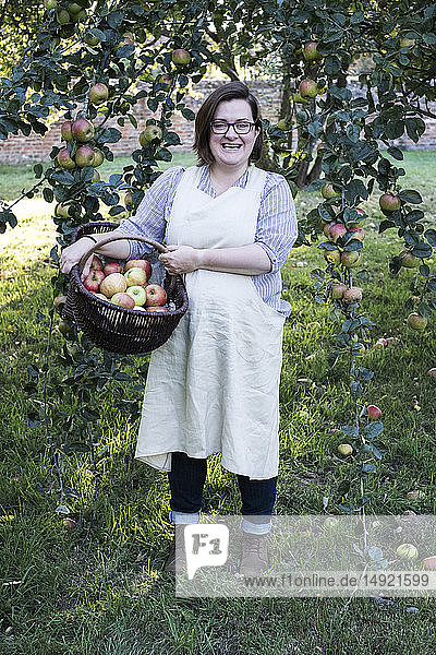 Frau mit Schürze hält braunen Weidenkorb mit frisch gepflückten Äpfeln und lächelt in die Kamera.