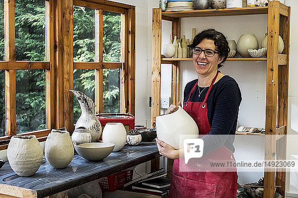 Frau mit roter Schürze steht in ihrer Werkstatt  hält eine Keramikvase und lächelt in die Kamera.