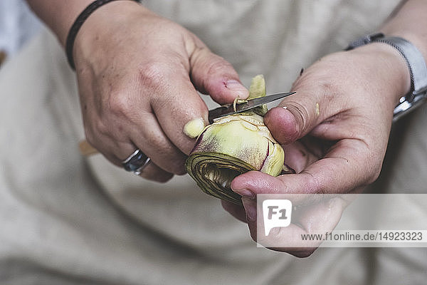 Close up of person peeling fresh artichoke with kitchen knife.