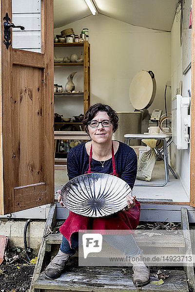 Woman wearing red apron sitting on steps outside her workshop  holding ceramic bowl with black line pattern.