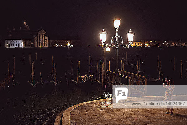 Woman taking picture of a streetlight by a lagoon in Venice  Veneto  Italy  at night.