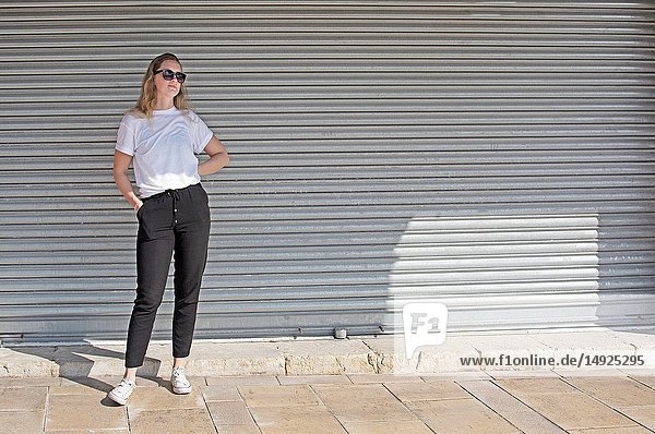 Full body photo of young casual sporty dressed blonde woman with sunglasses in white t-shirt against corrugated iron wall street style.