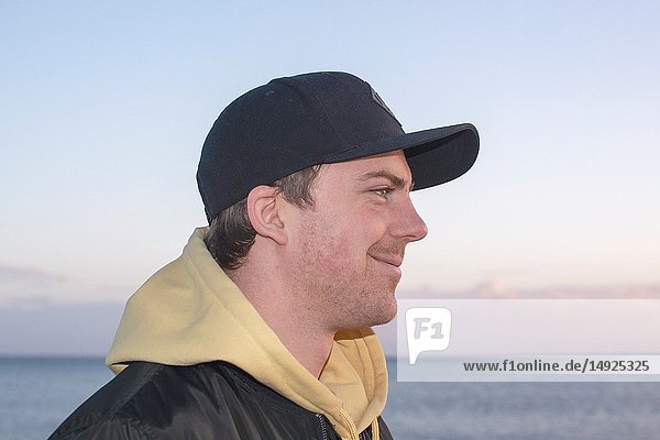 Profilaufnahme eines gut aussehenden  lächelnden  natürlich und lässig wirkenden Mannes in den späten Zwanzigern mit Kapuzenjacke und Mütze  der geradeaus blickt  mit dem Ozean als Horizont im Hintergrund.