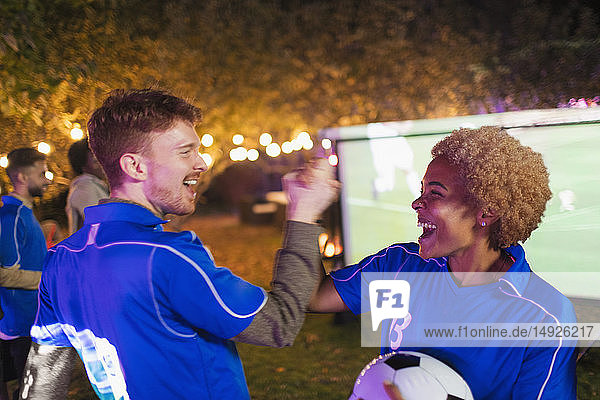 Glückliche Freunde  die jubelnd ein Fußballspiel auf einer Projektionsfläche im Hinterhof verfolgen