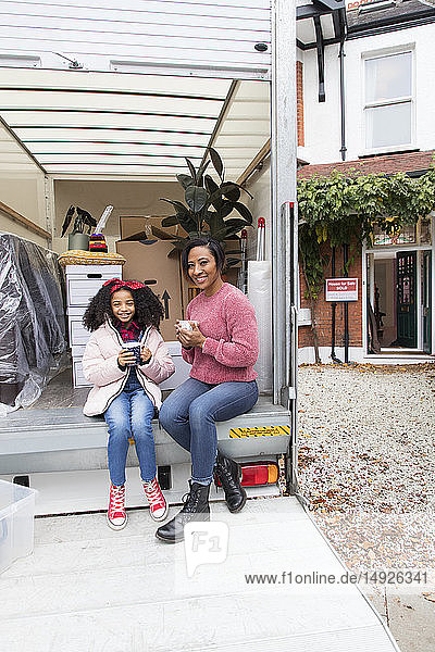 Porträt einer glücklichen Mutter und ihrer Tochter  die Tee trinken und eine Pause auf der Ladefläche eines Umzugswagens vor dem neuen Haus machen