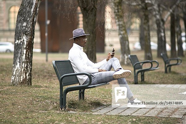 Stylish man sitting alone on bench in park  resting  using phone  looking aside  in city Munich  Germany.