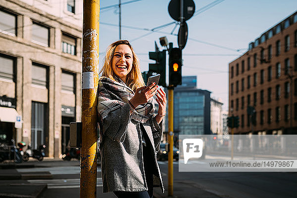 Junge Frau mit Smartphone lacht an Straßenbahnhaltestelle  Mailand  Italien