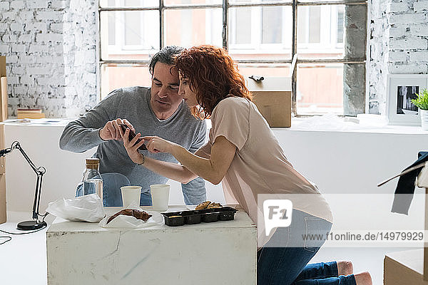 Couple moving into industrial style apartment kneeling using smartphone touchscreen