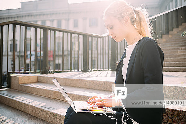 Junge Frau sitzt auf der Stadttreppe und tippt am Laptop