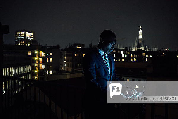 Junger Geschäftsmann tippt nachts auf dem Balkon seines Büros am Laptop