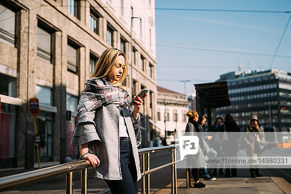 Junge Frau schaut auf Smartphone an der Straßenbahnhaltestelle