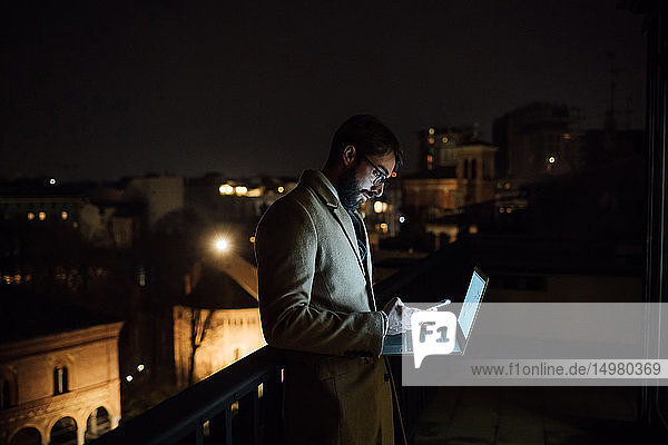 Mittlerer erwachsener Geschäftsmann schaut nachts auf dem Balkon seines Büros auf sein Smartphone