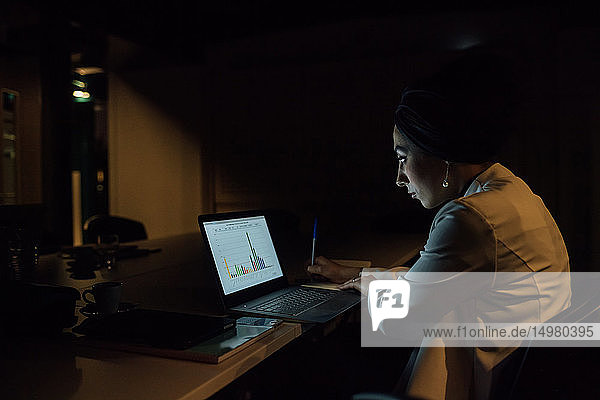 Junge Geschäftsfrau  die nachts im Büro sitzt und auf ihren Laptop schaut