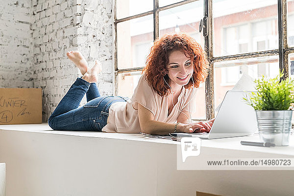 Mittelgroße erwachsene Frau, die in eine Wohnung im Industriestil zieht und auf der Fensterbank liegt und am Laptop tippt