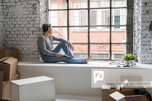 Mann zieht in eine Wohnung im Industriestil sitzt auf der Fensterbank und schaut durchs Fenster