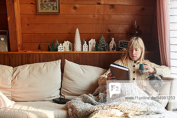 Woman reading book with dog on sofa at home