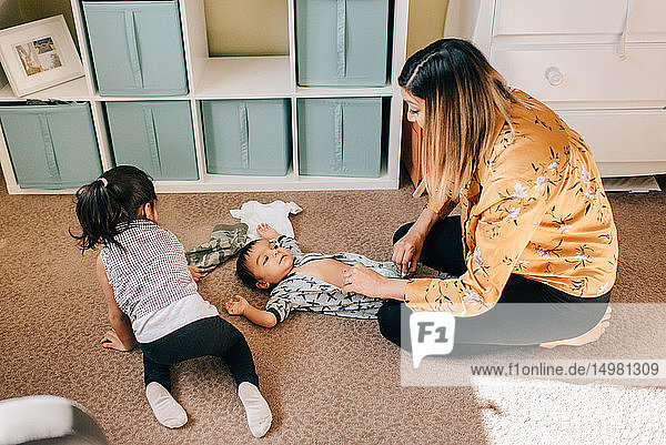 Mädchen krabbelt auf dem Kinderzimmerboden  während die Mutter den kleinen Bruder anzieht