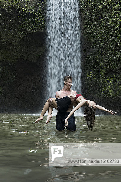 Junger Mann trägt junge Frau im Fluss am Tibumana-Wasserfall in Bali  Indonesien