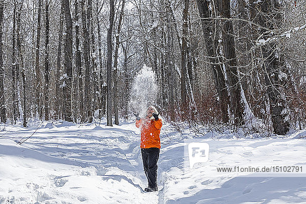 Ältere Frau in orangefarbenem Mantel wirft Schnee bei kahlen Bäumen