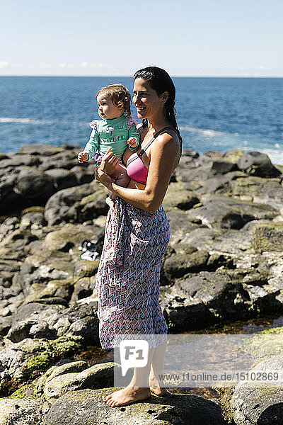 Woman holding baby daughter on rocks at beach