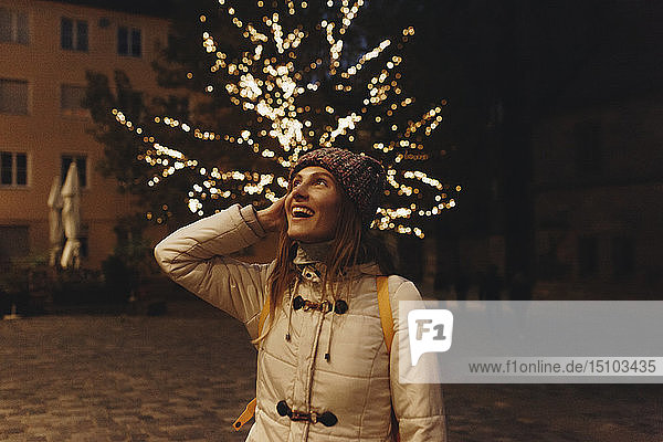 Lächelnde Frau mit Mantel bei Lichterketten im Baum bei Nacht