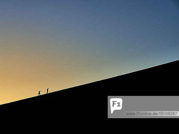 Silhouette von zwei Menschen  die bei Sonnenaufgang auf Big Daddy wandern  Namib-Wüste  Namibia