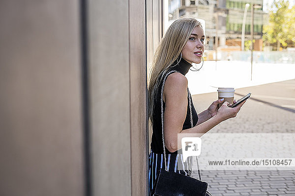 Junge blonde Frau mit Kaffee zum Mitnehmen und Handy an die Wand gelehnt und mit Abstand betrachtet
