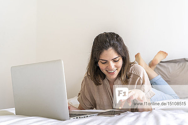 Glückliche junge Frau liegt auf dem Bett und benutzt Smartphone und Laptop