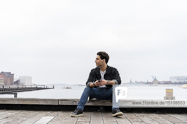 Dänemark  Kopenhagen  junger Mann sitzt mit Handy am Wasser