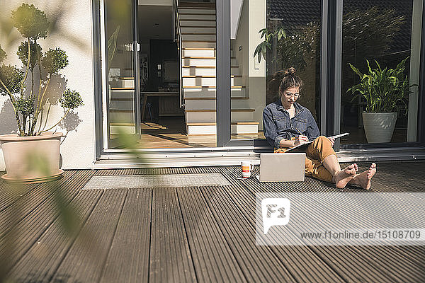 Junge Frau sitzt zu Hause auf der Terrasse und arbeitet mit Buch und Laptop