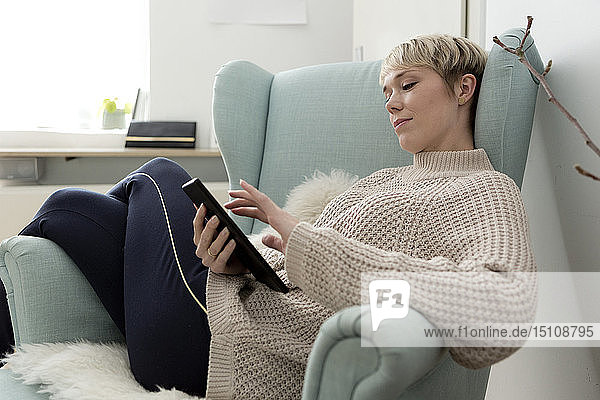 Entspannte Frau sitzt zu Hause im Sessel und benutzt Tablette