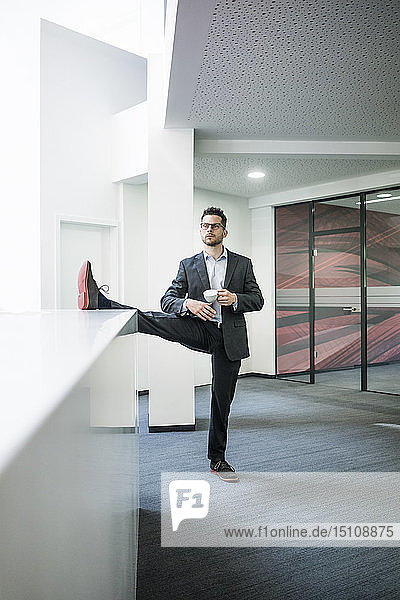 Businessman with cup of coffee standing on one leg in office