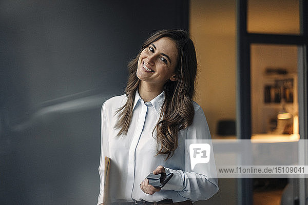 Porträt einer lächelnden jungen Geschäftsfrau  die einen Ordner und ein Mobiltelefon in der Hand hält