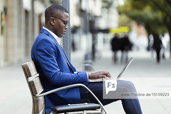Junger Geschäftsmann in blauer Anzugjacke sitzt auf Bank und benutzt Laptop