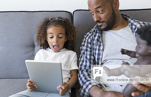 Vater und aufgeregte Tochter sitzen zu Hause auf der Couch und schauen auf die Tablette