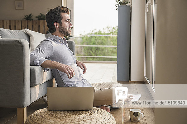 Junger Mann sitzt zu Hause auf dem Boden  benutzt einen Laptop und schaut aus dem Fenster