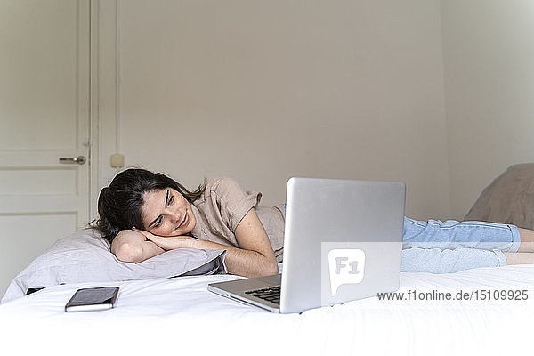 Lächelnde junge Frau auf dem Bett liegend mit Smartphone und Laptop