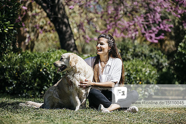 Happy woman sitting on meadow in city park with her Labrador Retriever enjoying sunlight