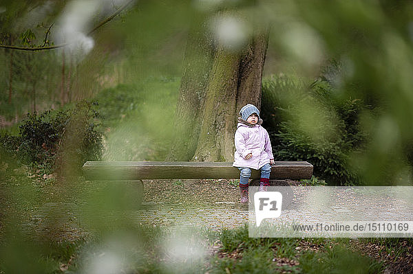 Kleines Mädchen mit blauem Hut und rosa Mantel auf einer Parkbank sitzend