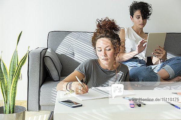 Junge Frau macht zu Hause Notizen  während ein Freund auf der Couch sitzt und ein Tablett benutzt