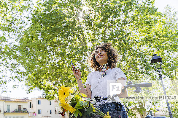 Lächelnde Frau mit Handy  Kopfhörern und Fahrrad im Park