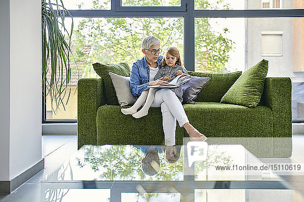 Großmutter sitzt mit ihrer Enkelin auf dem Sofa und liest gemeinsam ein Buch